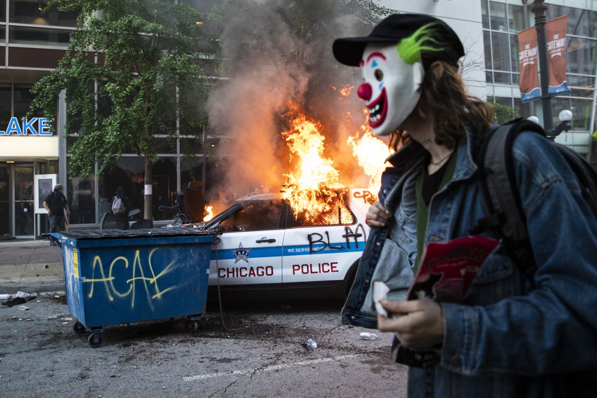 A man in a clown mask walks past a burning Chicago Police Department SUV near State and Lake on May 30, 2020.