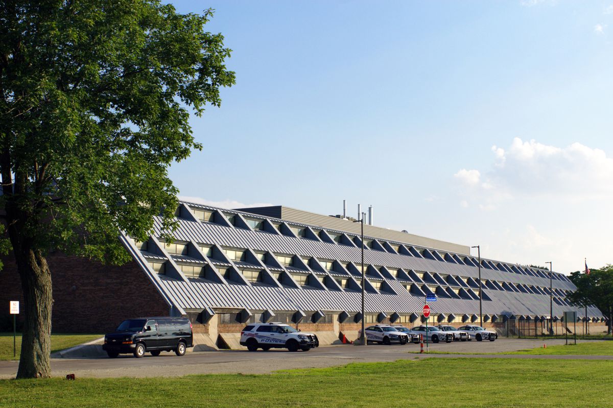 A long building with a slanted metal roof and skylights that stretches nearly to the ground. A few police cars sit in the parking lot.