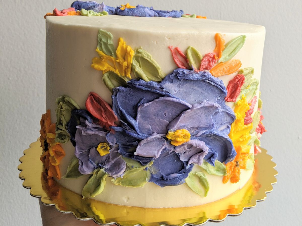 A cake with cream-colored frosting with large blue, yellow, and orange flowers on it.