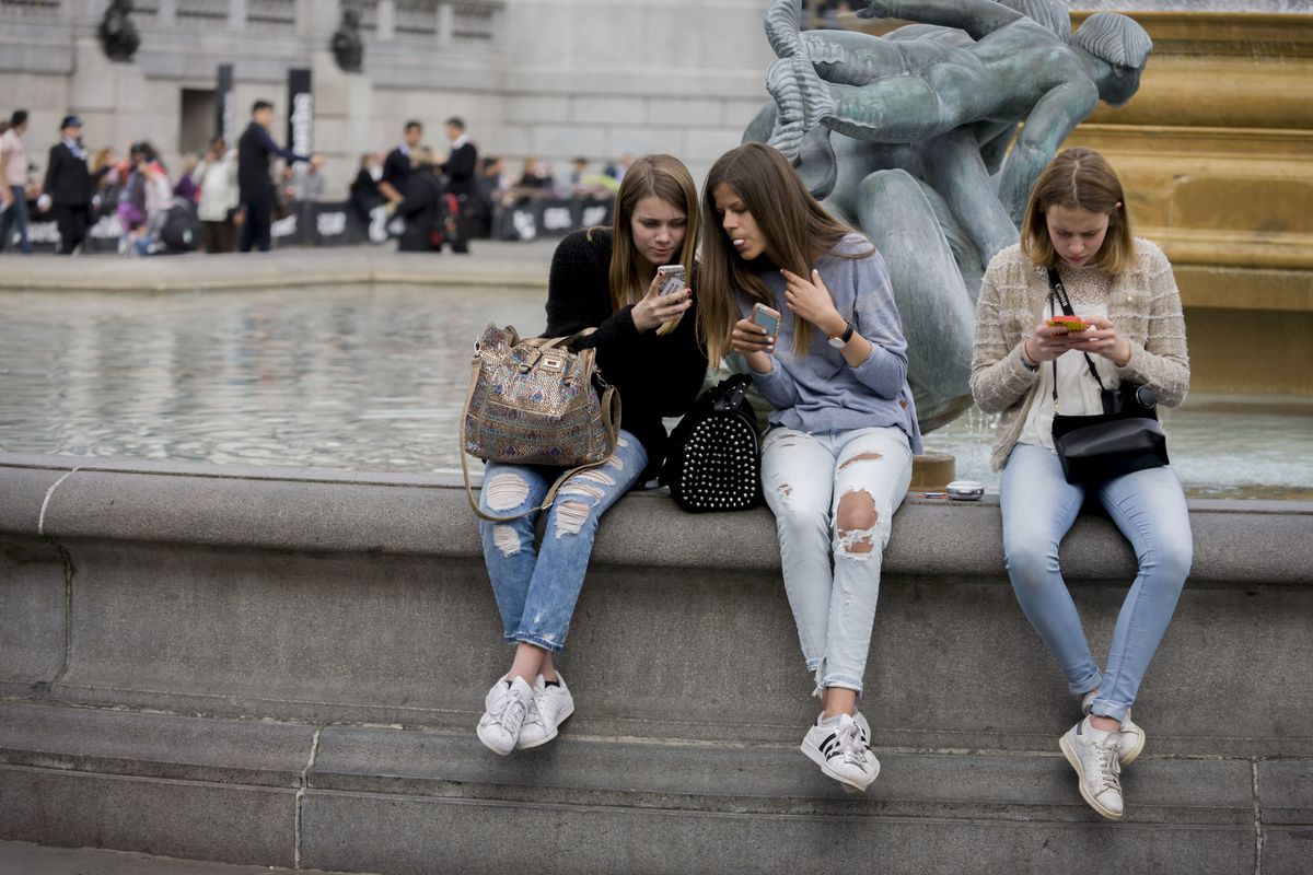Three teenage girls look at their phones while sitting beside a fountain in London.