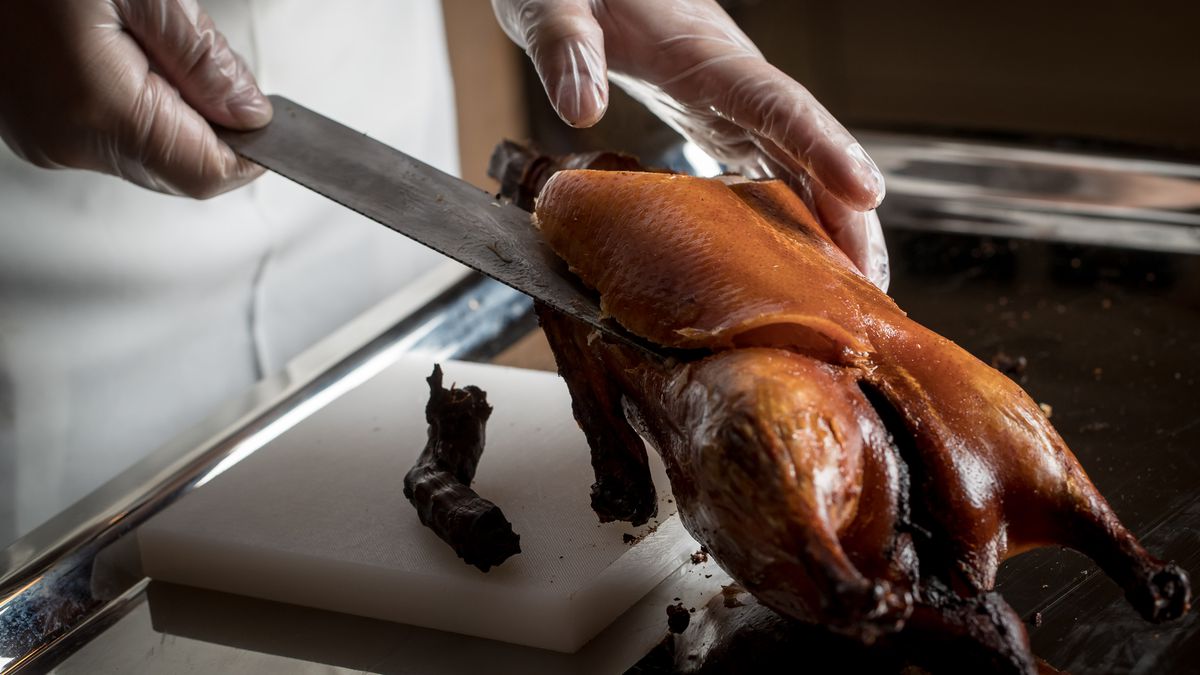 A chef’s gloved hands reaching towards a roast duck on a table. One hand holds a knife which is propping up the duck