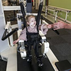 Chloe Caldwell, 3, smiles as she reaches for her dad's hand as she learns to walk again with the assistance of a Locomat machine at Neuroworx in Sandy on Friday, March 8, 2019. The machine helps individuals who have suffered paralysis of varying degrees to learn to walk again by supporting their full weight at first and decreasing the support as they regain strength. Caldwell attends therapy at Neuroworx five times a week after suffering an injury to her spinal cord during a heart operation.