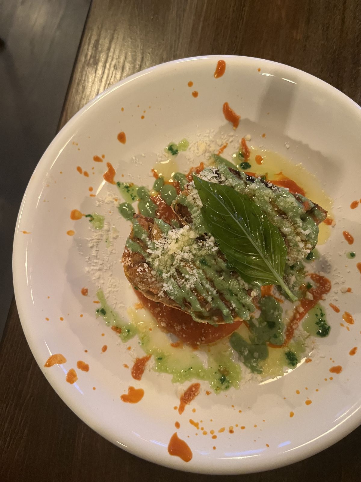 A baked eggplant dish with green and orange sauces splattered around the plate and a green basil leaf set on top.