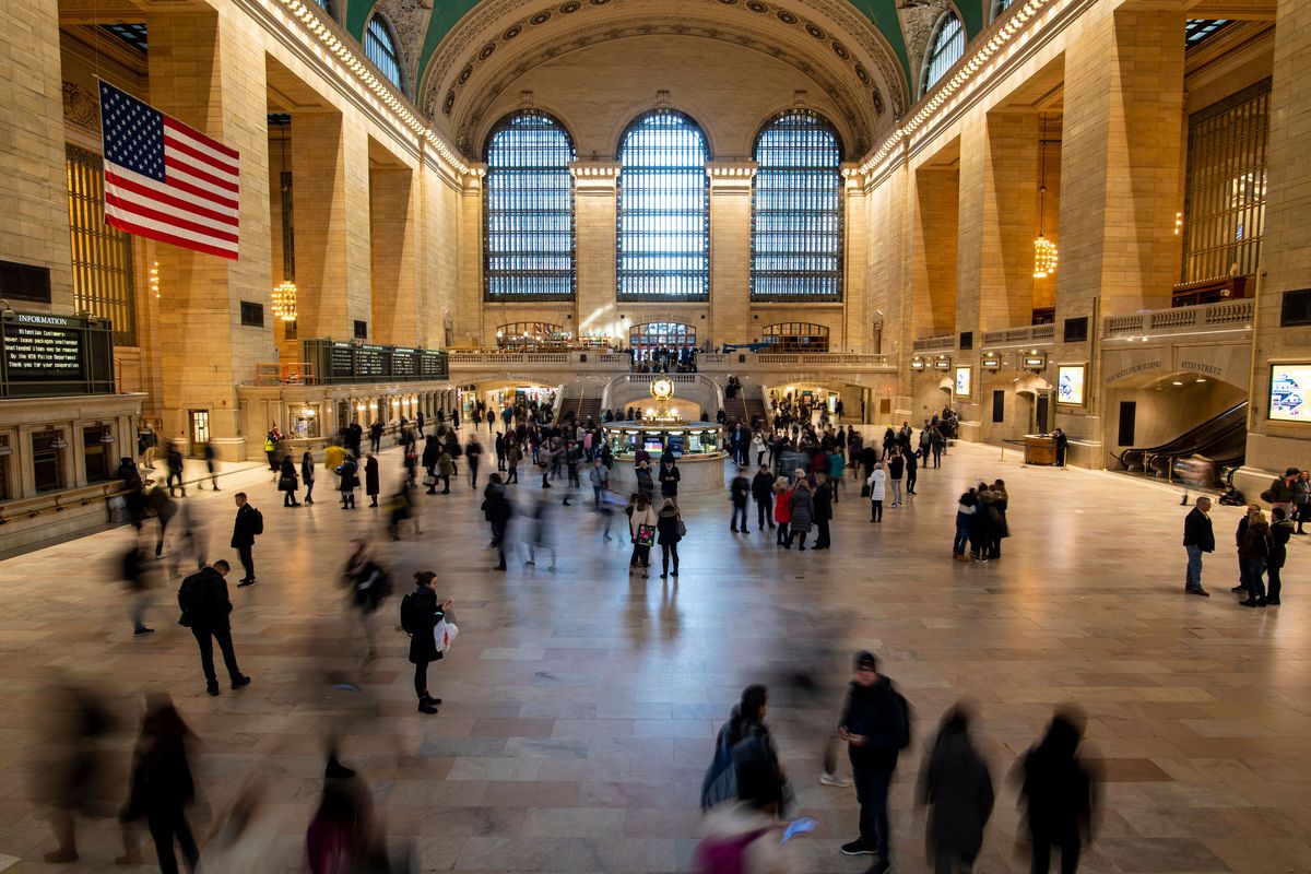 A high-ceilinged interior of a train station with large windows and crowds of people walking across the main concourse. A large American flag hangs on the left wall.