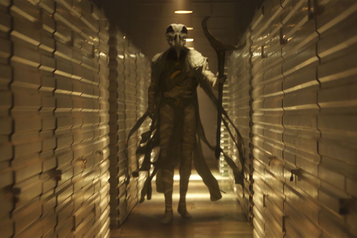 Konshu strides down a dark hallway in Moon Knight. He is a figure in mummy wrappings and a robe, carrying a huge staff with a crescent moon at the end of it. His head is the skull of a huge bird, floating above his shoulders.