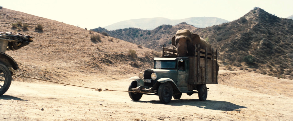 An elephant in a truck being dragged up a dirt hill by another car offscreen in Babylon