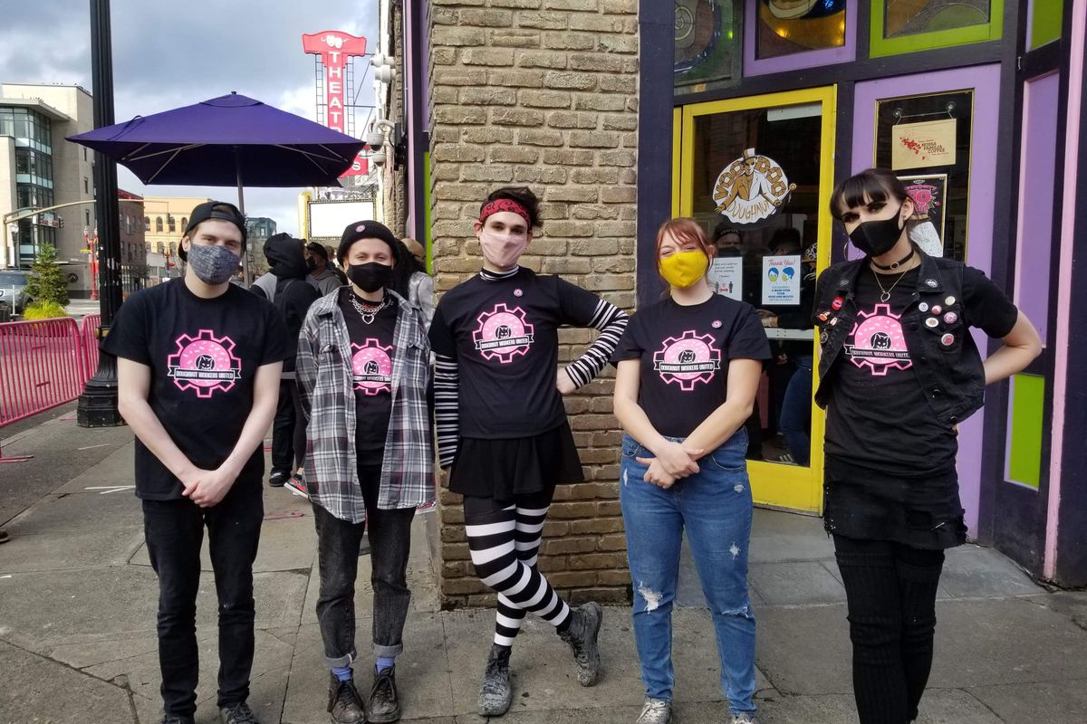 Five employees of Voodoo Doughnut wearing the signature black and pink t-shirts stand outside the door of the shop’s downtown location