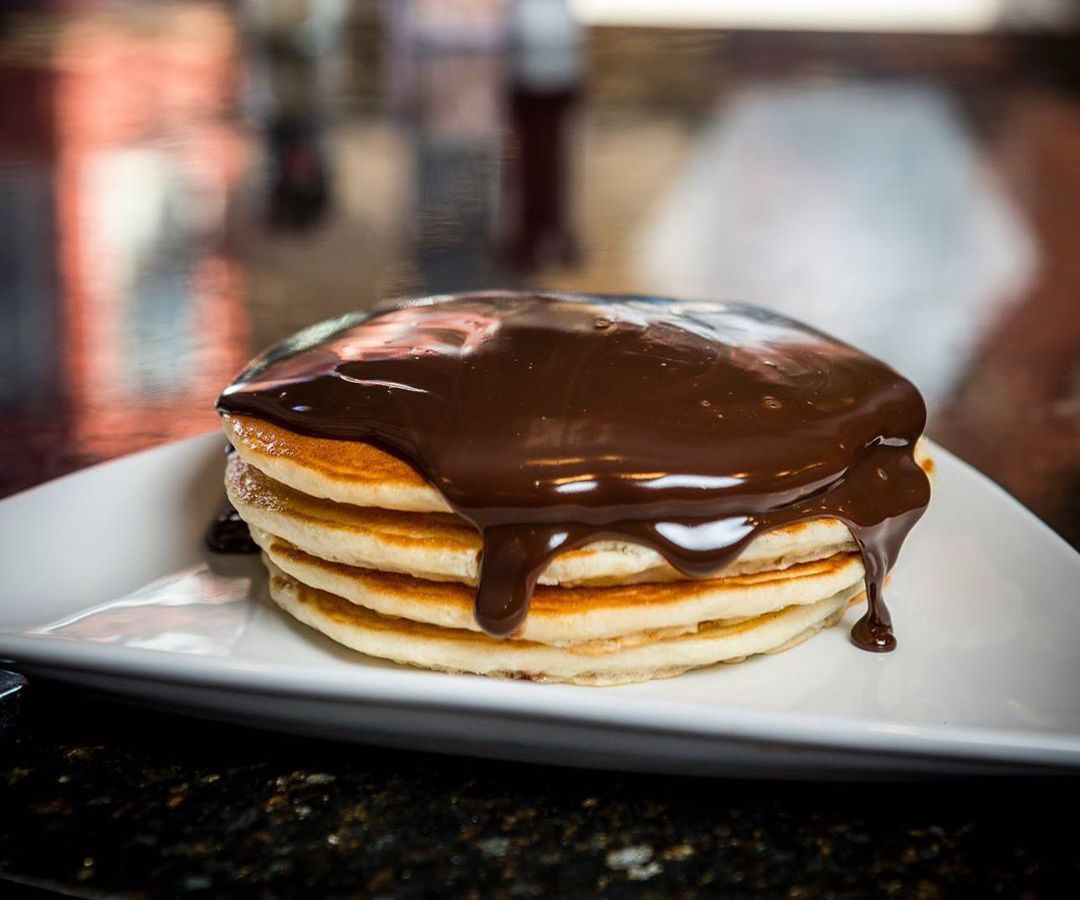 A stack of thin pancakes are covered with a thick, shiny, dripping layer of chocolate ganache
