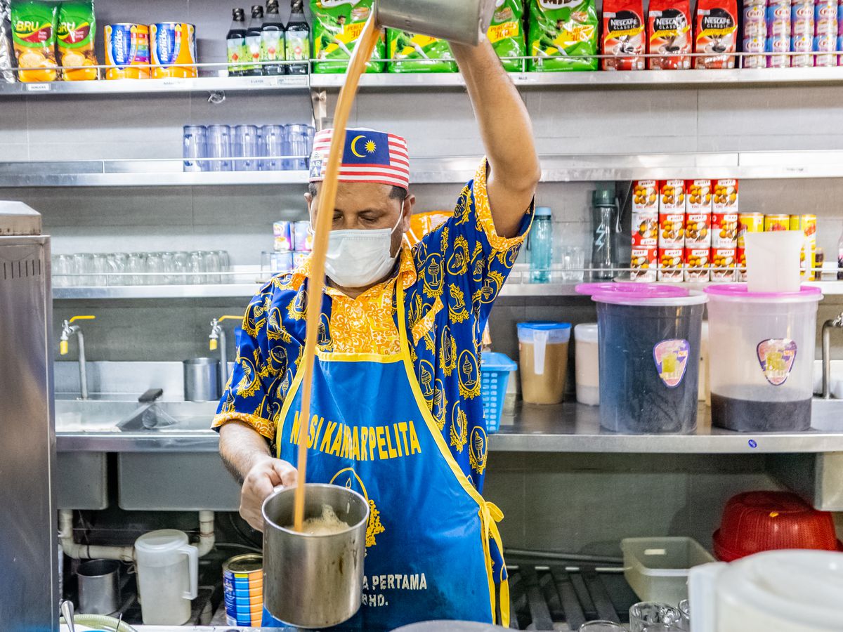 A cook, wearing a branded apron and Malaysian flag hat, pours tea from a metal pitcher to a pot.