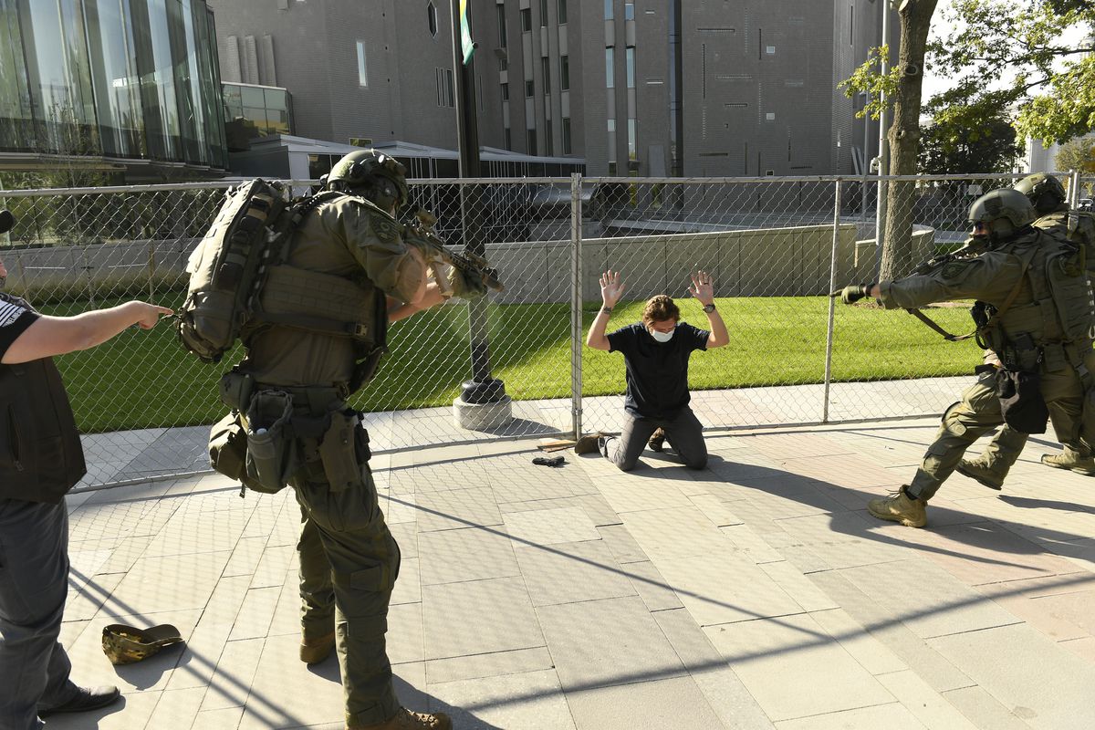 A white man, in a black T-shirt, black jeans, and a white mask, kneels in front of a chain link fence with his hands up. Beside him is a handgun. Three law enforcement officers in military-style body armor, including helmets, surround him, their assault rifles pointed at him. A man in the corner of the photo in a black T-shirt points at the suspect.