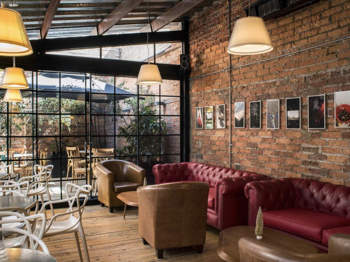 A restaurant interior with exposed brick wall, glass panel ceiling and wall, leather couches, and tables. 