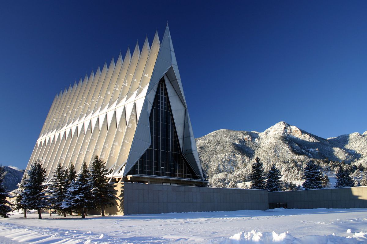 The exterior of the Cadet Chapel in Colorado. The facade consists of multiple spires and colorful stained glass. There are mountains and trees in the background. The ground is covered with snow.