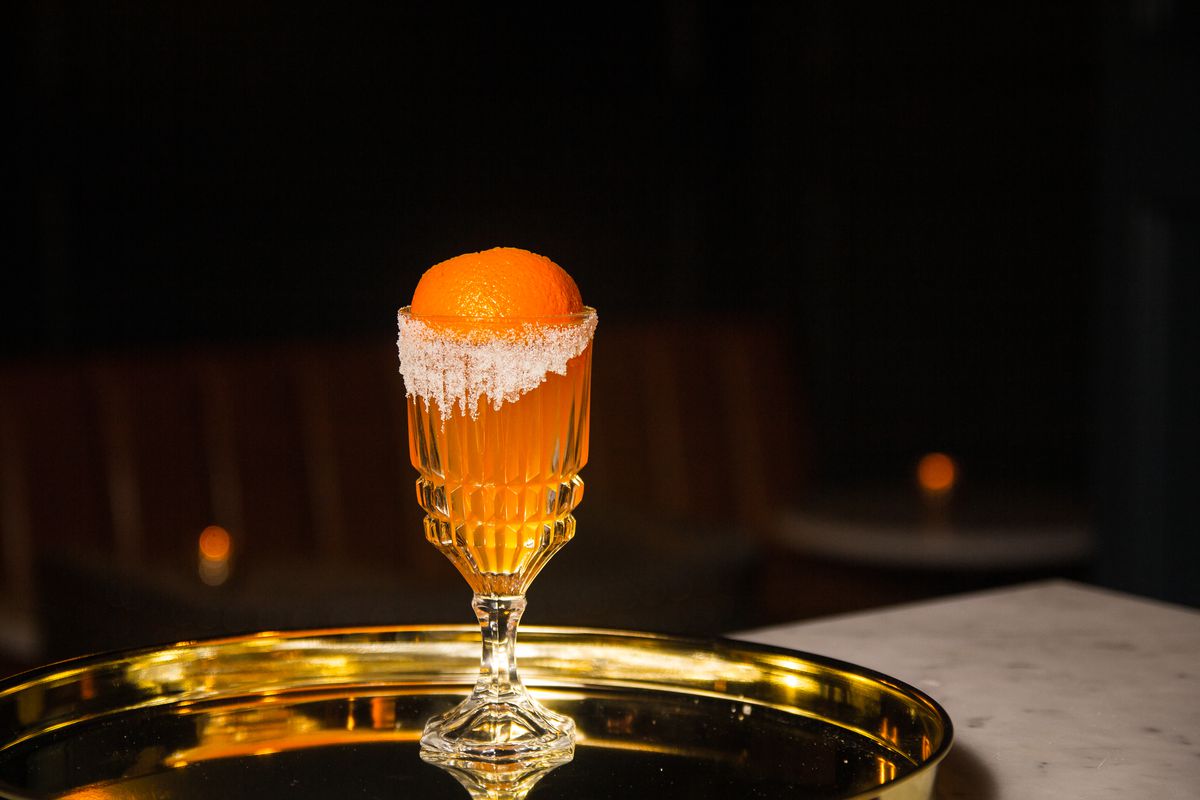 A cocktail in a tall glass with an orange sphere on top and white dust along the rim of the glass, all on a tray.