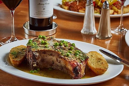 A plate of pork chop vesuvio on a table with a glass of red wine.