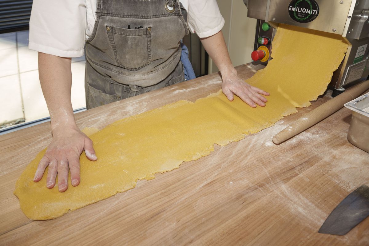 A thick sheet of pasta with rough edges protruding from a metal pasta maker, and a person in a gray apron and white shirt placing their hands on top of it.