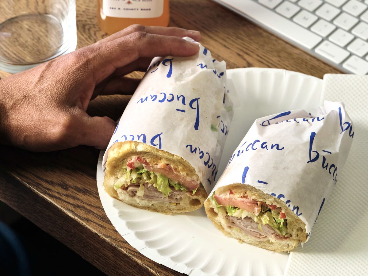 In the foreground of this picture is a sandwich, cut in half and wrapped in white wax paper with Buccan Sandwich Shop printed on the paper. The sandwich is and Italian sandwich with deli meats and provolone cheese. There is a man’s hand to the left of the picture, just touching the sandwich. In the background is a glass, an orange bottled drink and the keypad of a computer.