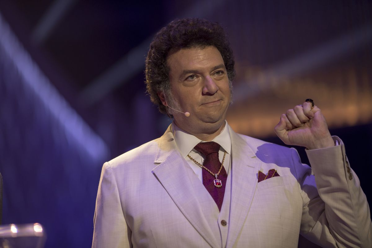 Danny McBride plays Jesse Gemstone wearing a suit, a headset microphone, and holding up his fist.