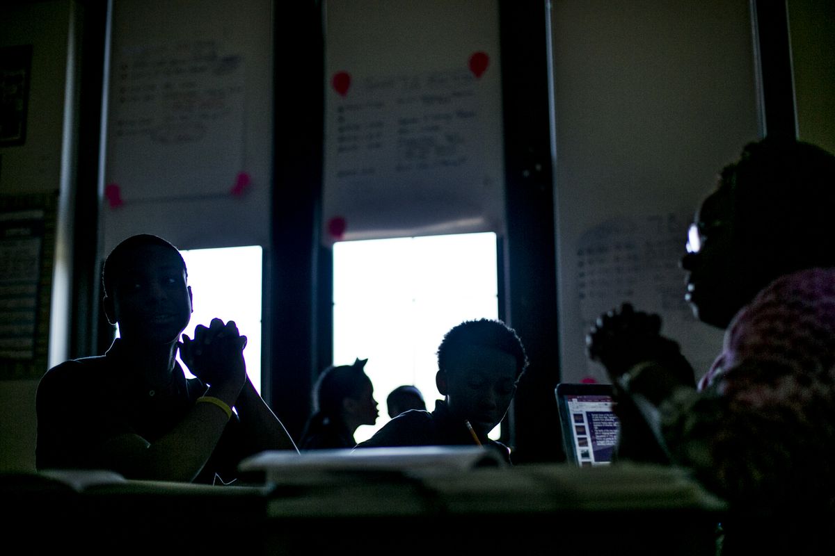 Students and their teacher sit in a dark classroom, the students silhouetted against the windows.