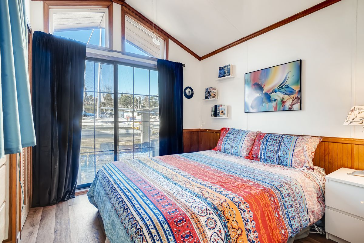 A bedroom in the rear of the has a quilted queen bed, a window with blue curtains, and vaulted ceilings. 