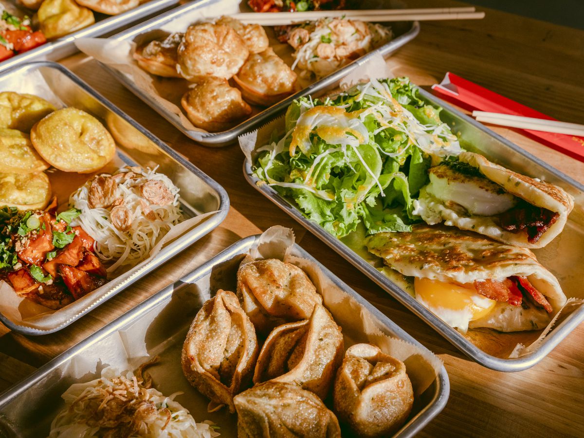 Metal trays filled with dumplings, egg sandwiches with scallion pancakes, and side salads, sitting on a wooden table in the sunlight.