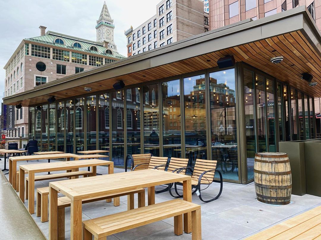 A glass-walled taproom is surrounded with a patio covered with light wooden picnic tables. Historical city buildings are visible in the background.