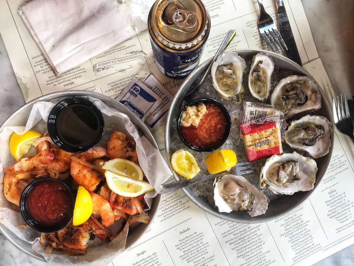 A half-dozen opened oysters atop ice on a tin platter with lemon wedges and cocktail sauce sice next to another tin plate will of cooked shrimp.