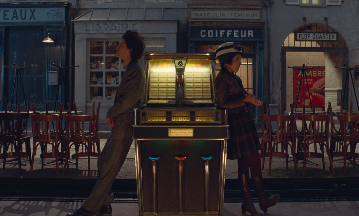 Timothée Chalamet and Lyna Khoudri lean on opposite sides of an outdoor jukebox (that’s a thing?), facing away from each other in Wes Anderson’s The French Dispatch.