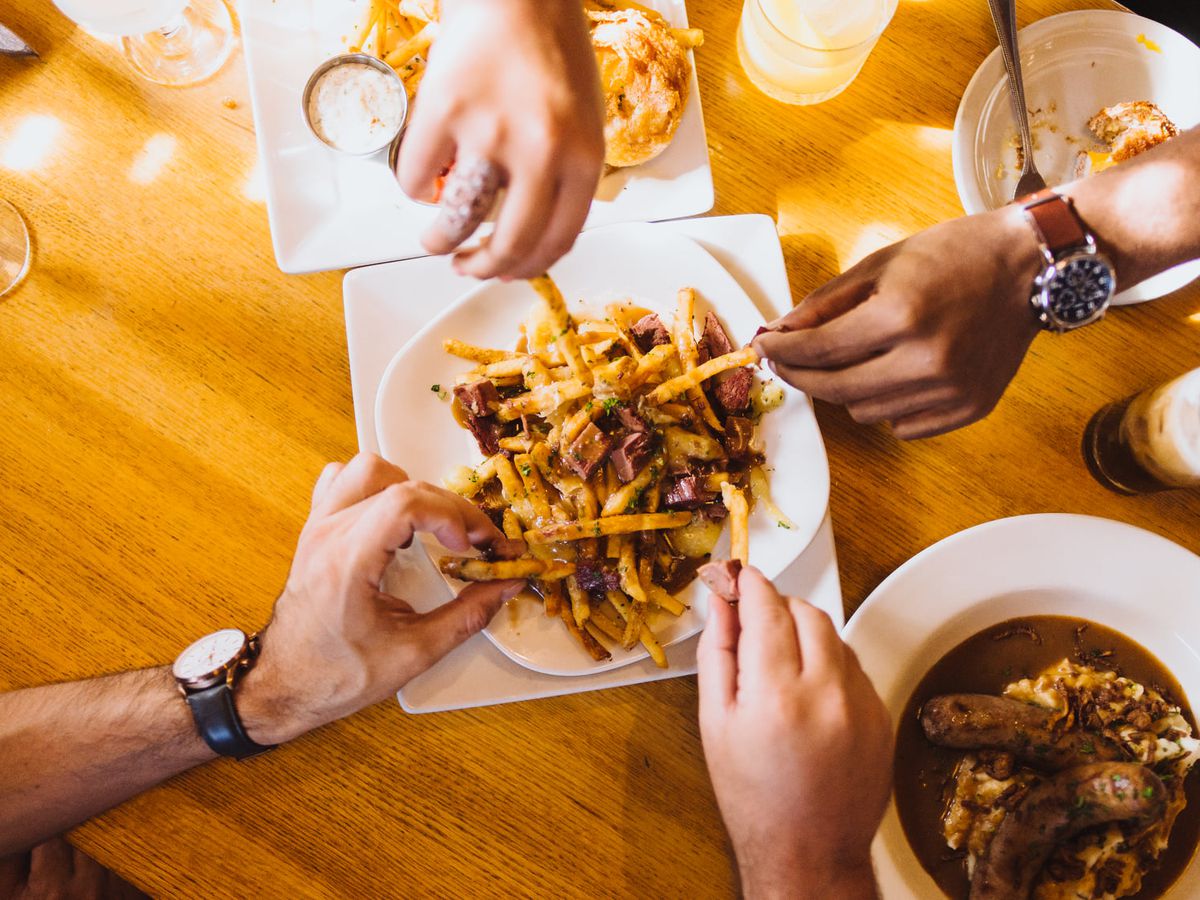 Hands reach toward a plate of fries covered in gravy and corned beef. A bowl of sausages and potatoes sits next to the pile of fries, as well as a bowl of mac and cheese.