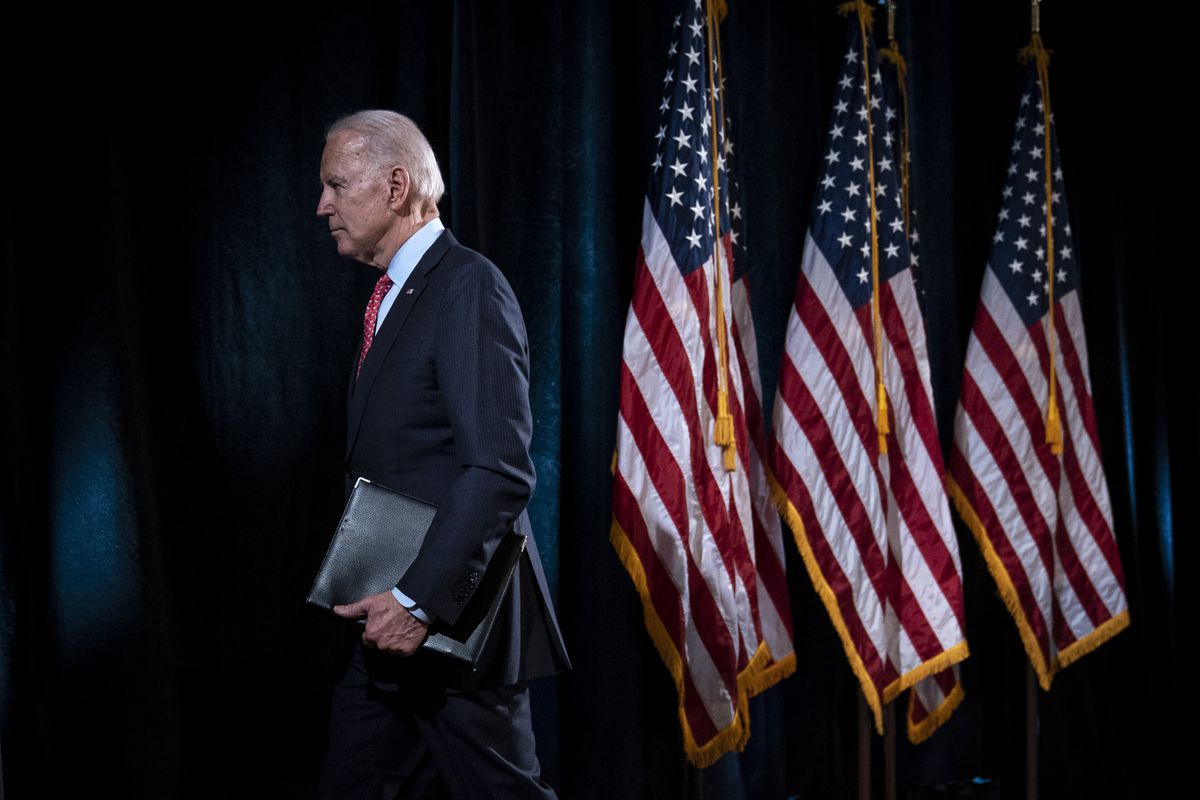 Biden clutches a leather binder as he walks past three US flags, while wearing a dark suit.