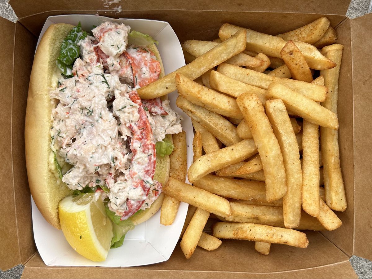 An overhead photograph of a takeout container with an oversized lobster roll and a pile of fries.