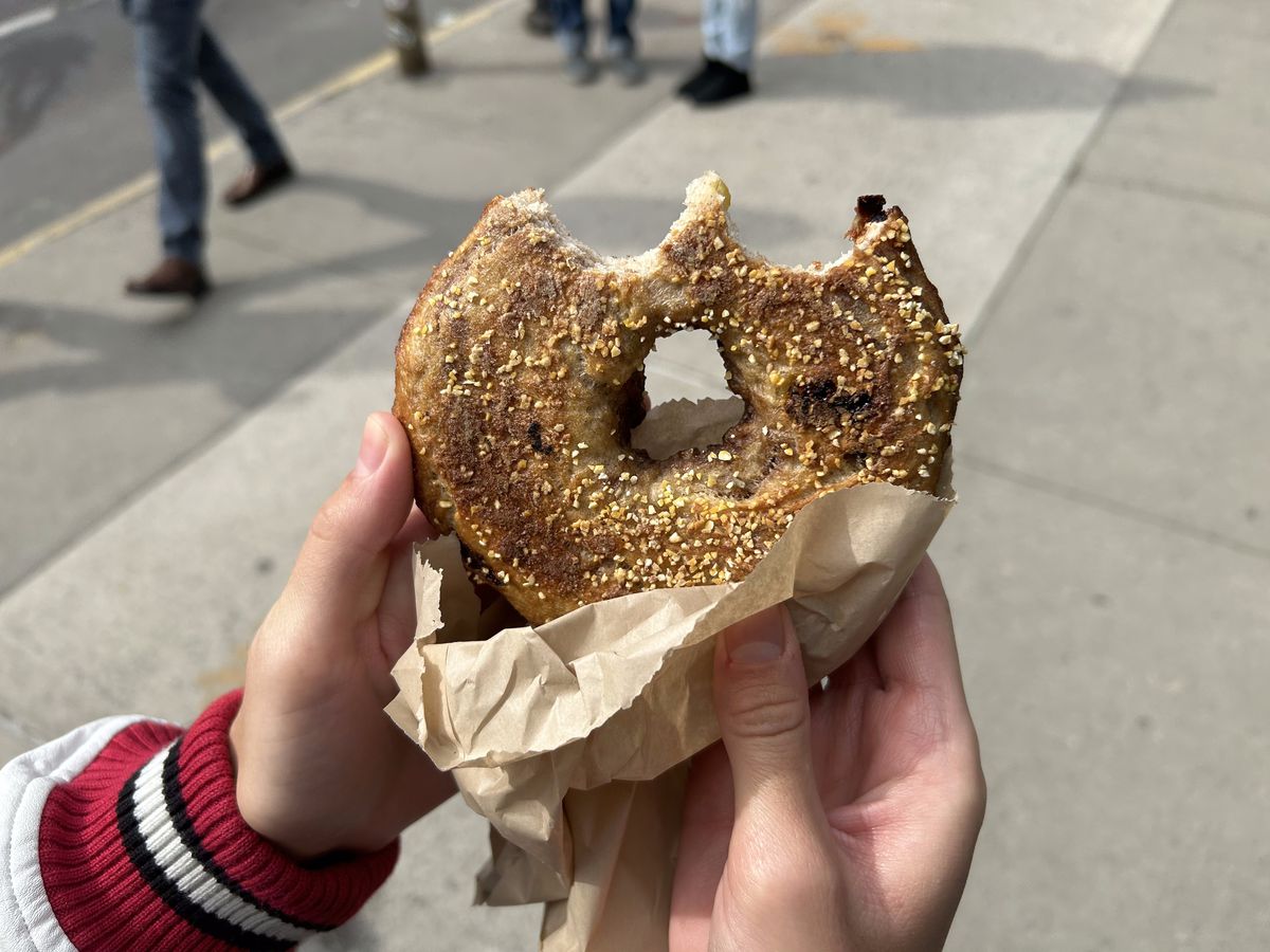 A hand holds a bitten bagel studded with pieces of corn meal on a sunny day.