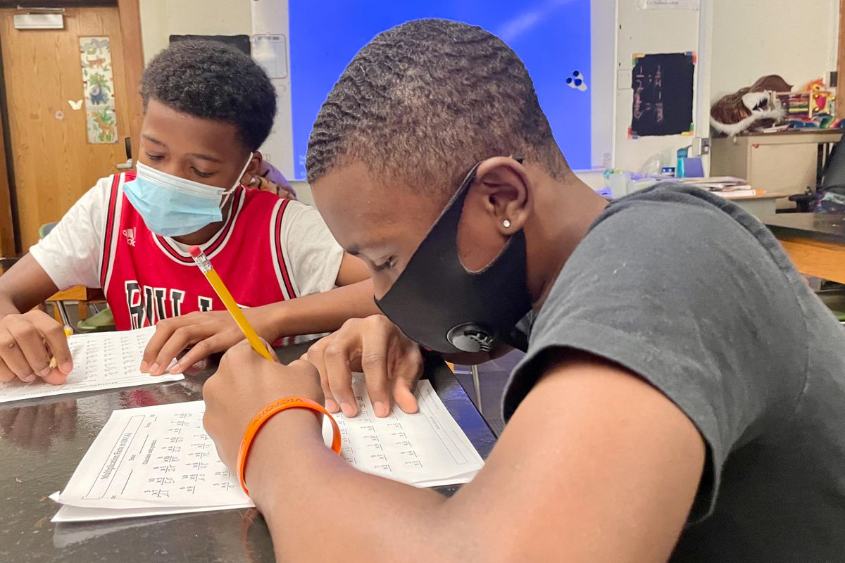 Two male middle school students work on mathematics worksheets at their desk. Both are wearing face masks.