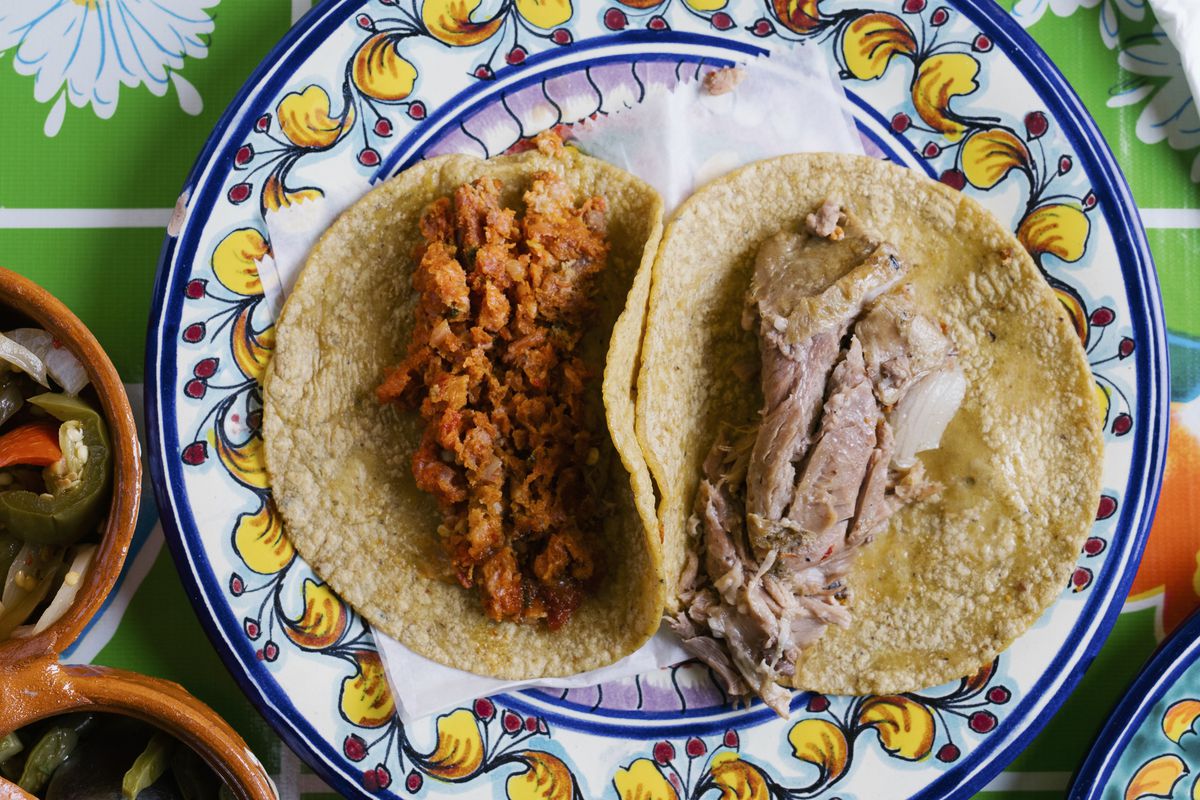 An overhead photograph of two corn tortillas, one filled with crumbly orange meat and another with brown- and pink-colored meat slices.