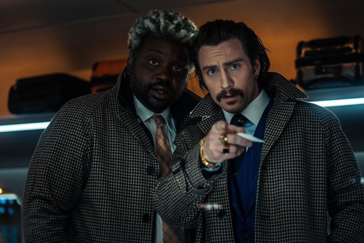 Lemon, played by Brian Tyree Henry, and Tangerine, played by Aaron Taylor-Johnson, are two slickly dressed assassins wearing blue suits under checked pea coats and are mugging directly at the camera, while Tangerine points at the viewer in the movie Bullet Train.