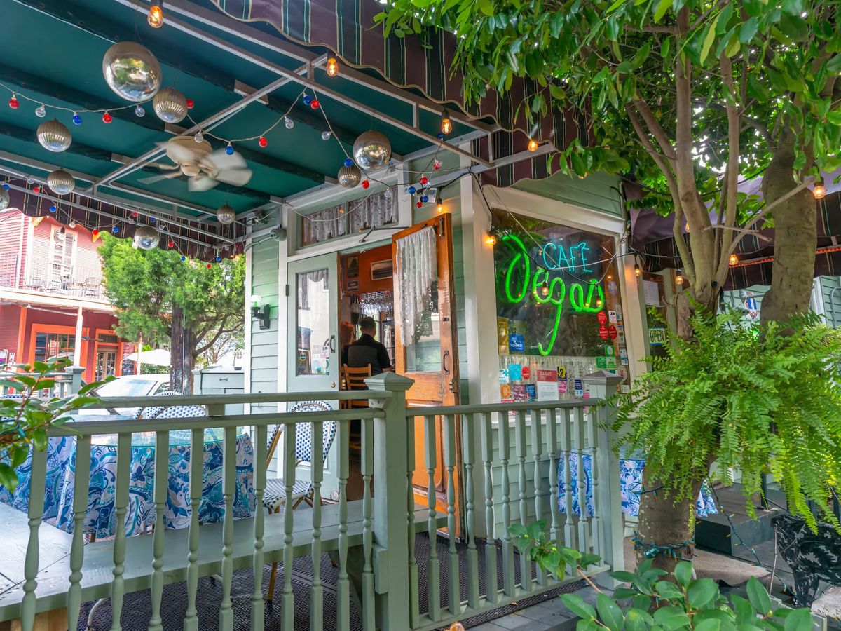 View of a tree-draped front porch and door opening into a restaurant with a neon Cafe Degas sign in the window. 