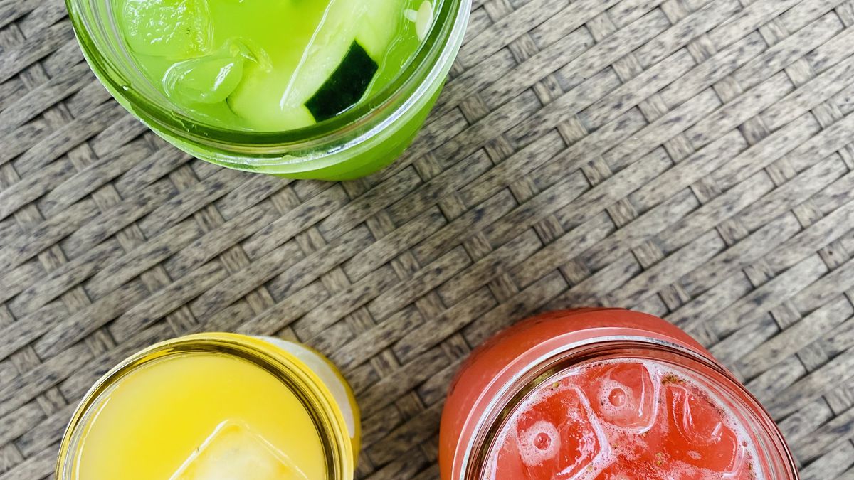 Three glasses of aguas frescas in red, green, and yellow on a brown plastic table.