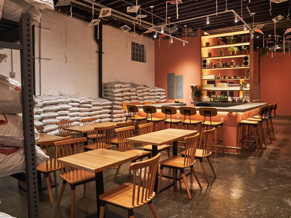 A high-ceilinged restaurant and bar is outfitted with a combination of tables and high-top chairs. Stacked bags of corn line its walls.