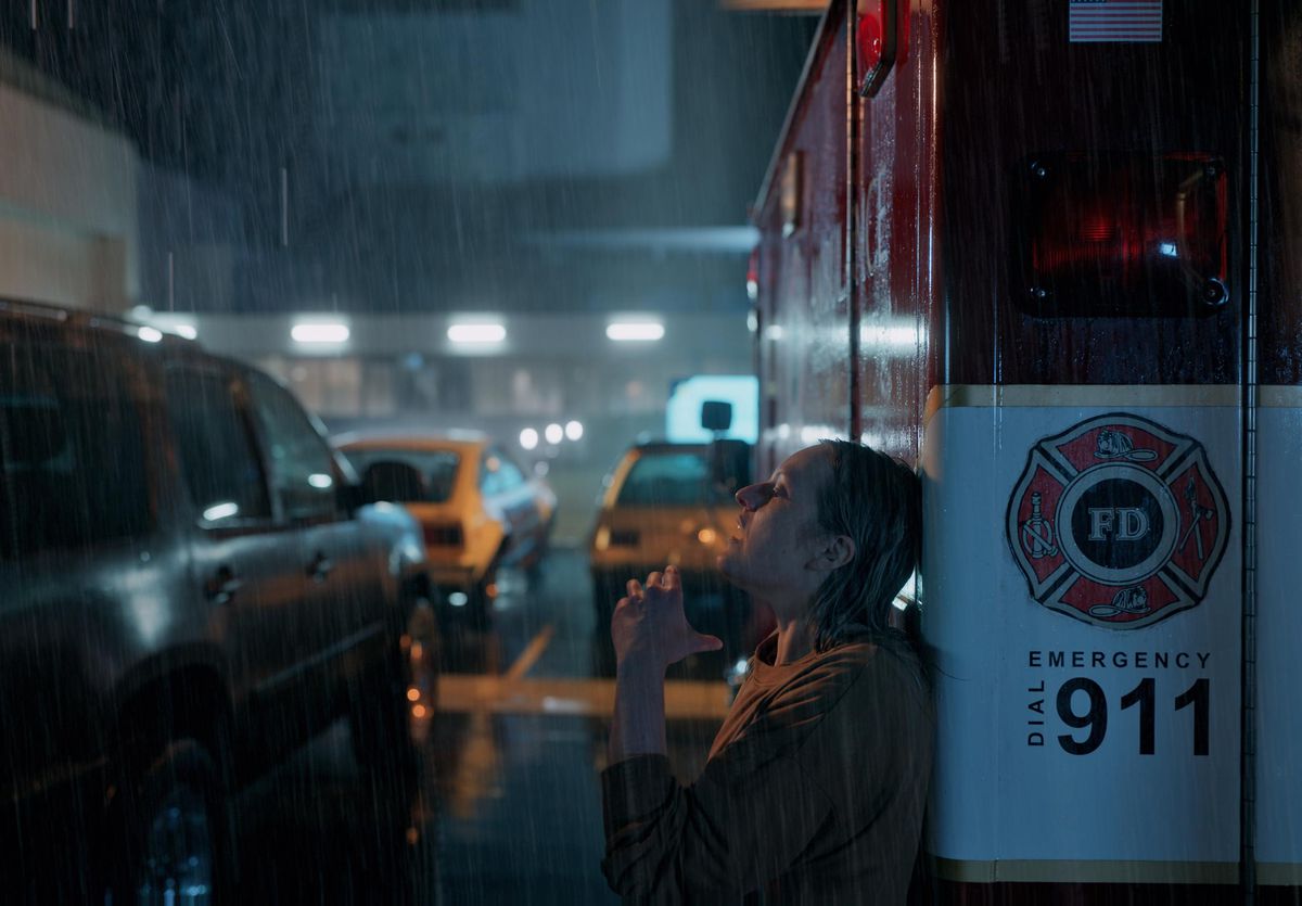 A blonde woman in a soaked beige sweater slumps against a fire department vehicle in the rain at night, her eyes closed and her mouth open as she appears to gasp for air.