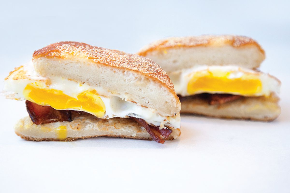 An eggy breakfast sandwich from Mike &amp; Patty’s, topped with bacon, sits on a white background