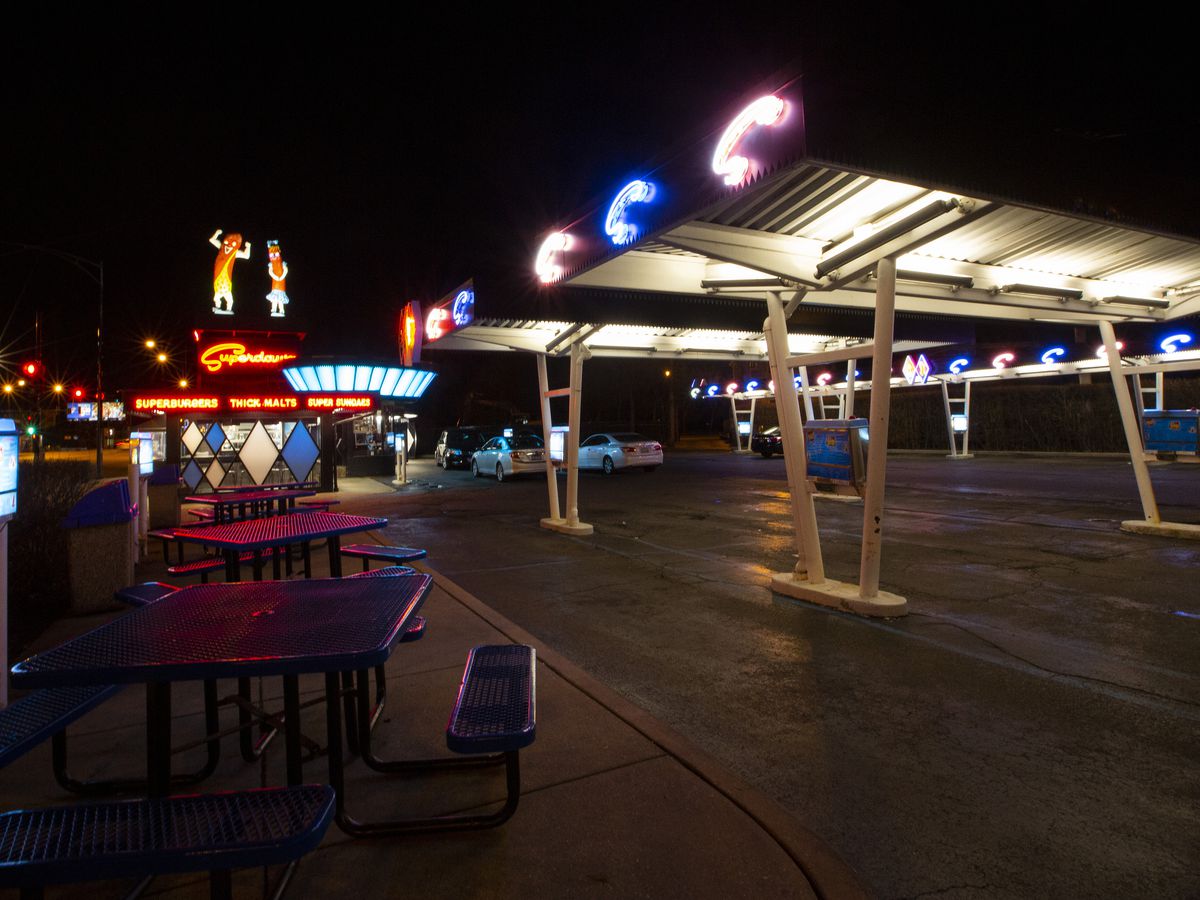 A drive in restaurant’s parking lot at night with two giant fiberglass wiener’s and plenty of neon.