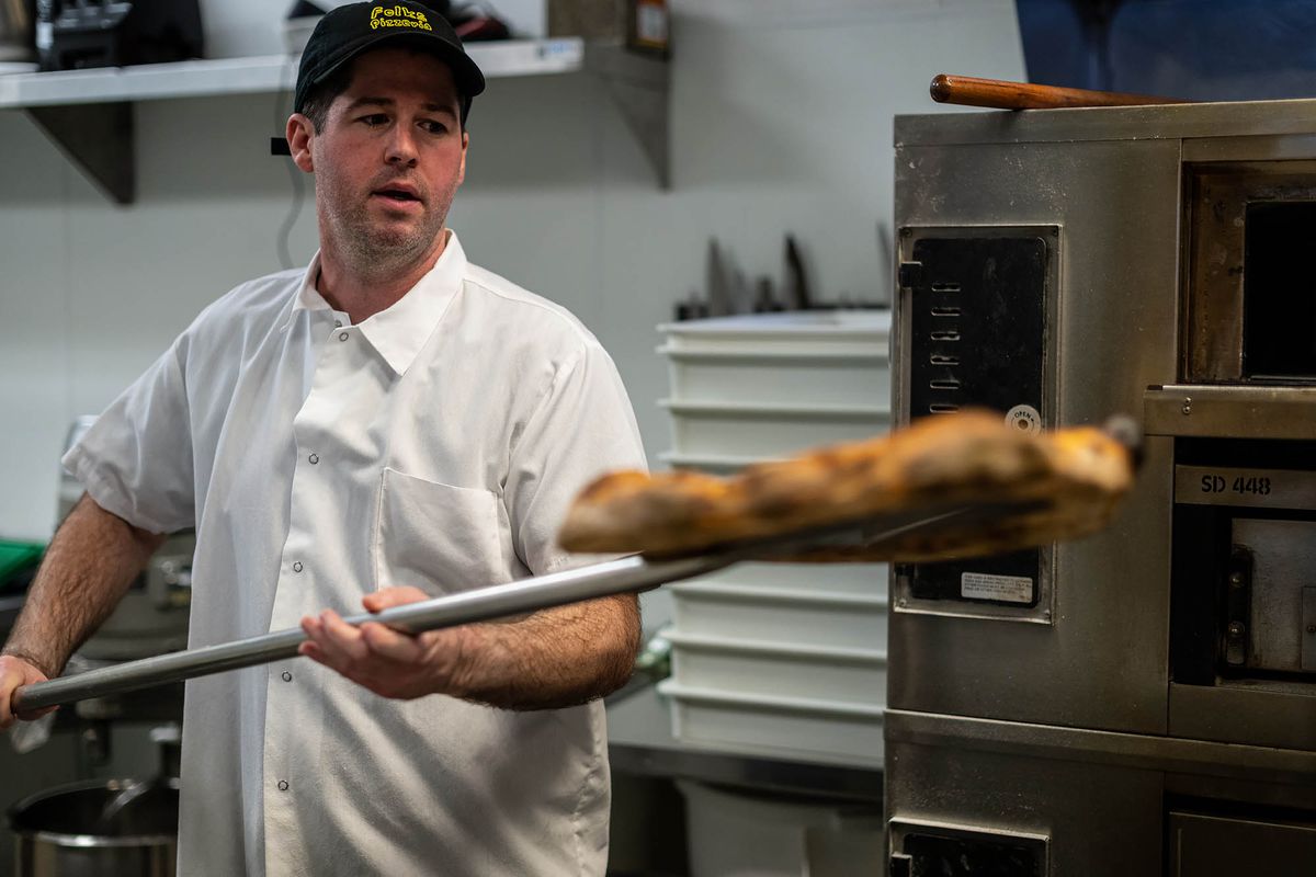 A chef in a black hat pulls out a burnished pizza using a long metal tool at a restaurant.