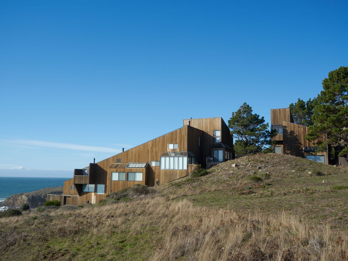A house on a cliff adjacent to a body of water. The house facade is brown and the house structure is angled.