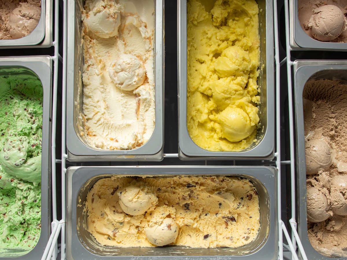 A case of seven different ice cream flavors lined up in metal bins, in green, yellow, and cream colors. 