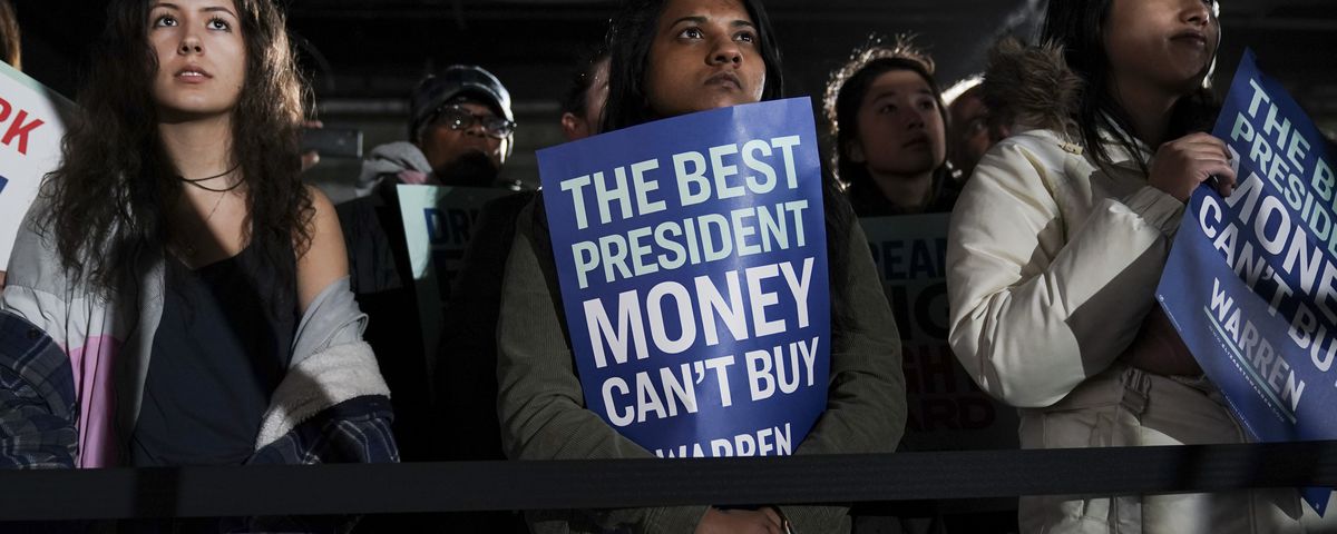 Elizabeth Warren fans stand at a barricade holding signs that read, “The best president money can’t buy.”