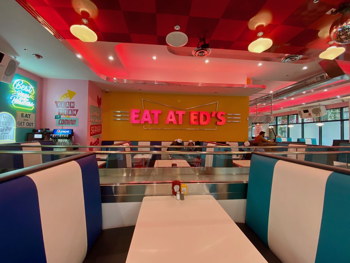 blue and white striped diner booths in the foreground in front of a yellow wall with a pink neon sign that reads “Eat at Ed’s”