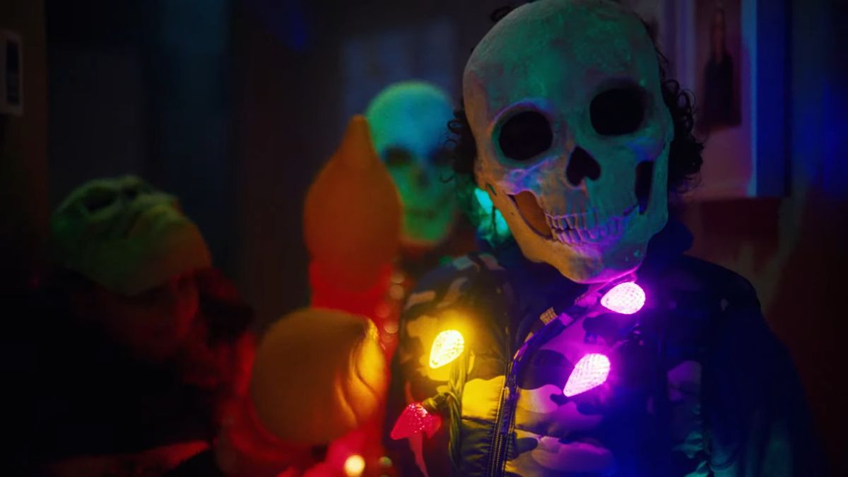 A person wearing a skull-faced mask and an elaborate costume with multi-colored lights strewn around their neck.