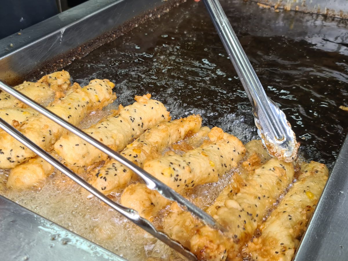Battered bananas frying in a large vat of oil with tongs hanging above.