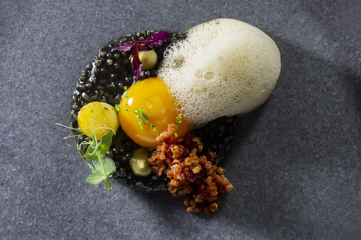A dish composed of a foam, egg yolk, caviar, and what appear to be bacon bits