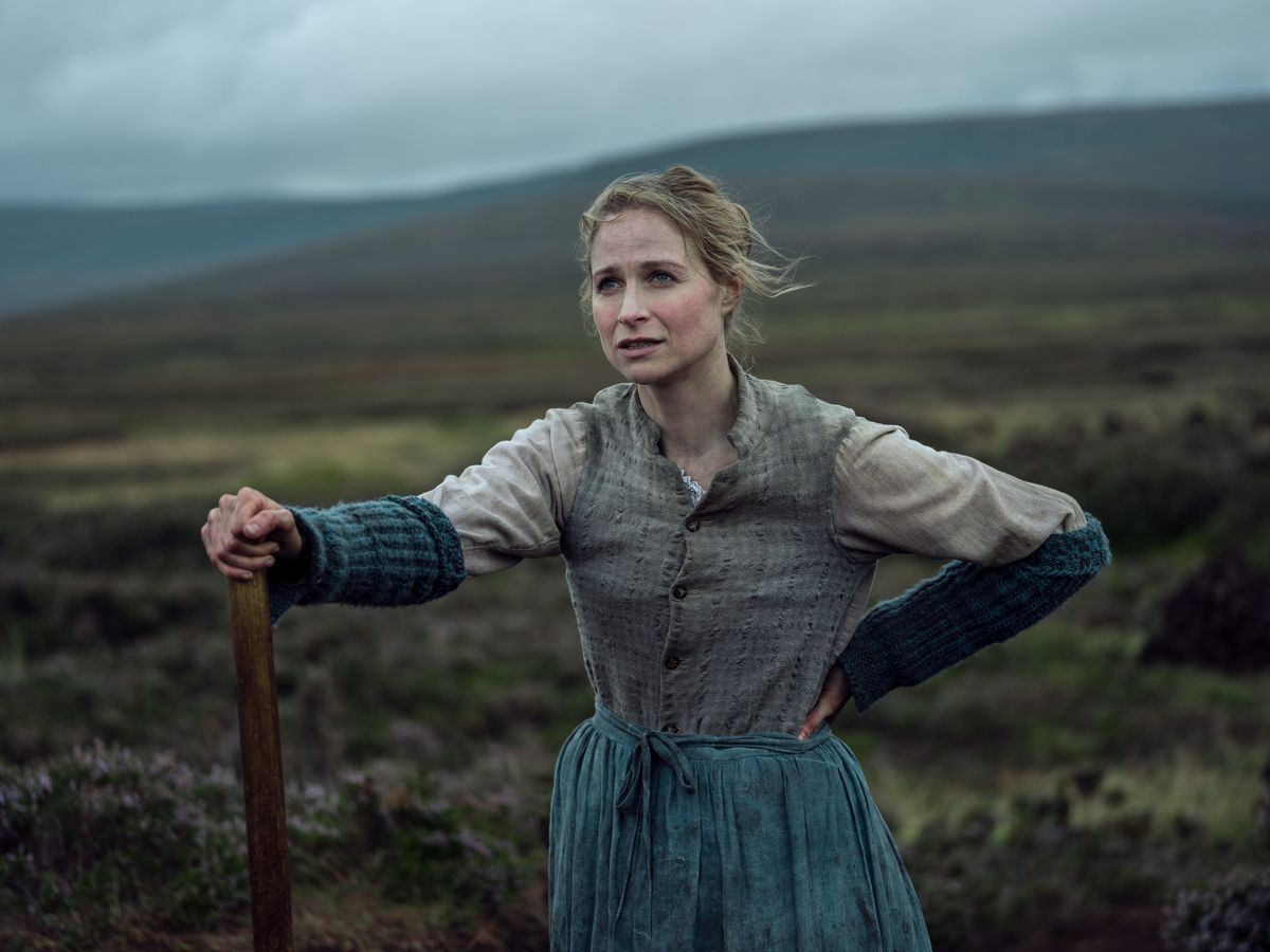 Kitty O’Donnell (Niamh Algar), a fair-skinned, blonde woman in a dirty blue skirt, stands in a field with her hands on a farm tool in The Wonder