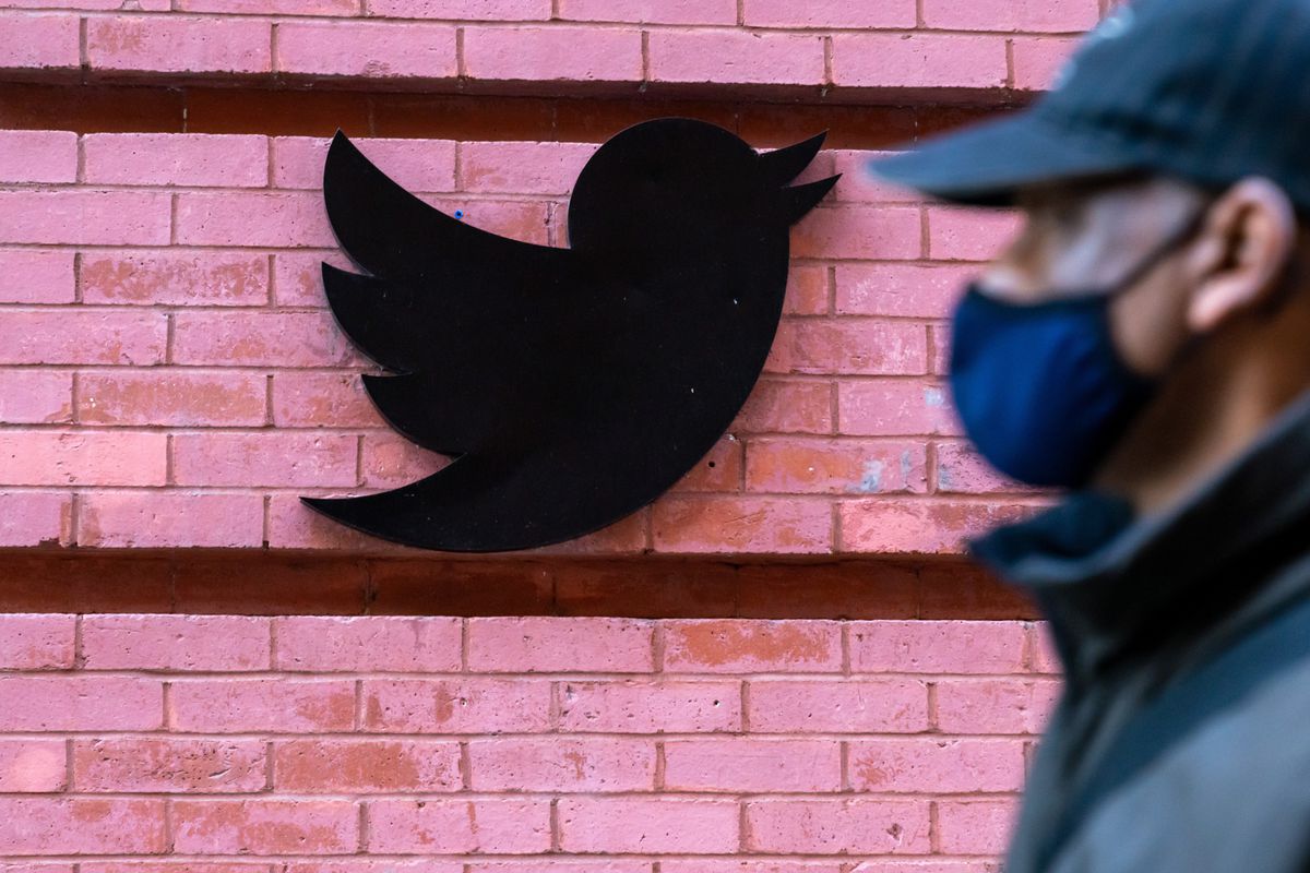 A man wearing a face mask walks past a Twitter logo on the side of a building.